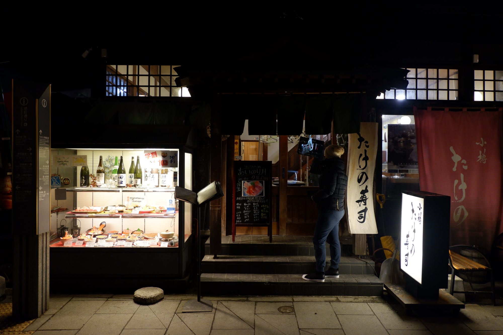 ผู้หญิงยืนอยู่หน้าร้านซูชิ "ทาเคโนะซูชิ" ในตอนกลางคืน มีเมนูภาษาอังกฤษและป้าย "ทาเคโนะซูชิ
