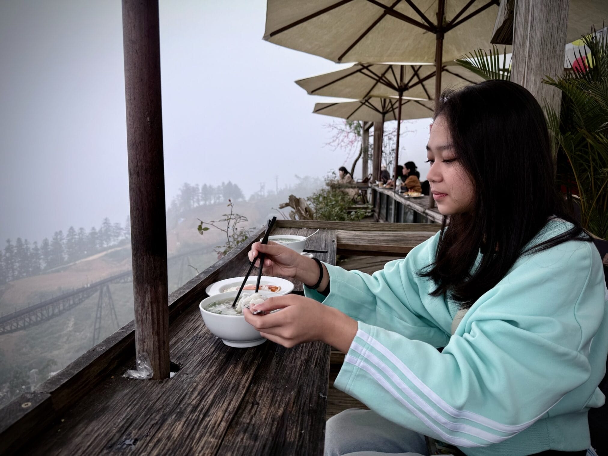 ผู้หญิงเอเชียทานก๋วยเตี๋ยวบนระเบียงพร้อมวิวทิวทัศน์ภูเขาที่ปกคลุมไปด้วยหมอก