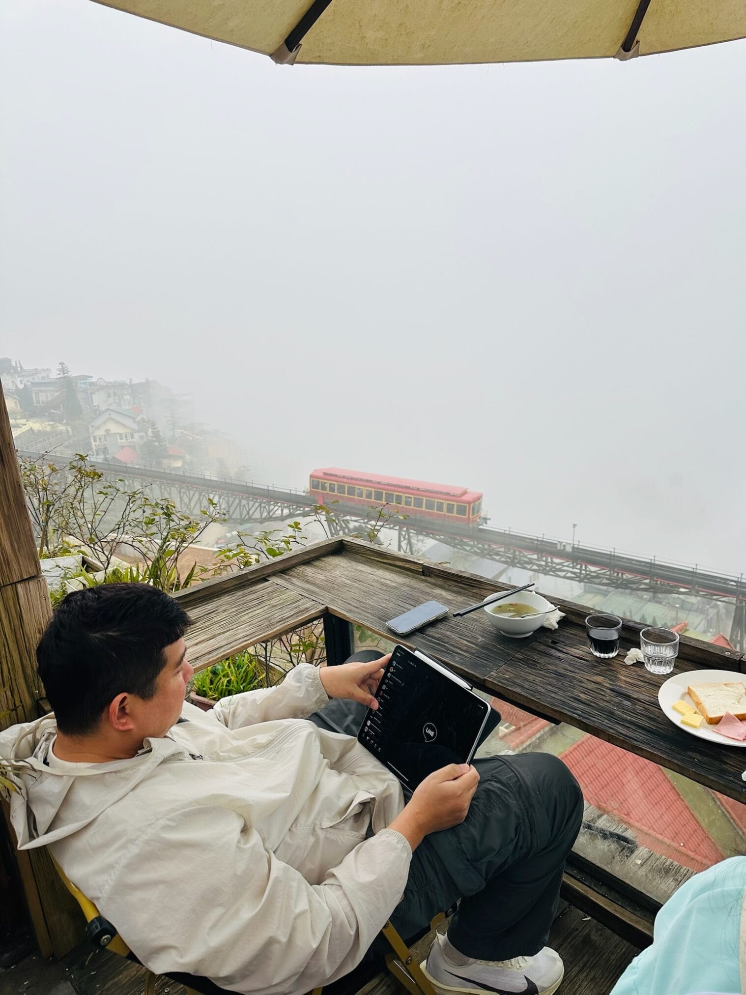 ชายกำลังดูแท็บเล็ตที่ระเบียง มีรถไฟเก่าสีแดงวิ่งผ่านในสายหมอก