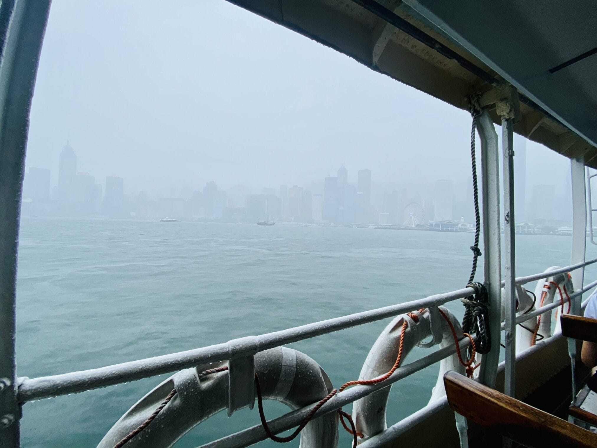 วิวจากเรือเฟอร์รี่ฮ่องกงในวันที่ฝนตก มองเห็นตึกและชิงช้าสวรรค์รางๆ