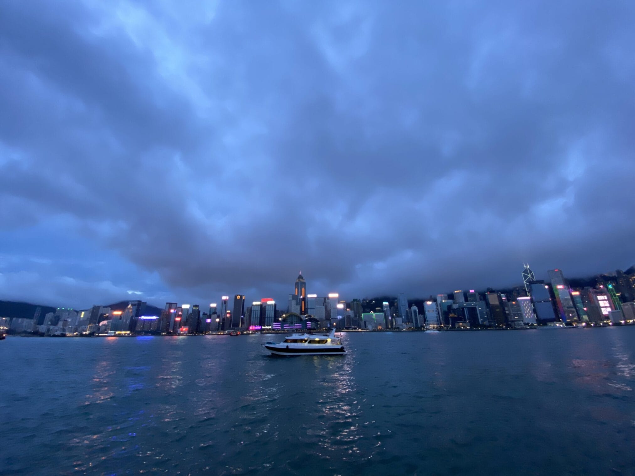 วิวเมืองฮ่องกงยามค่ำคืน มีเรือลำหนึ่งลอยอยู่บนอ่าวและท้องฟ้ามีเมฆหนา Hong Kong night cityscape with boat on harbor and cloudy sky