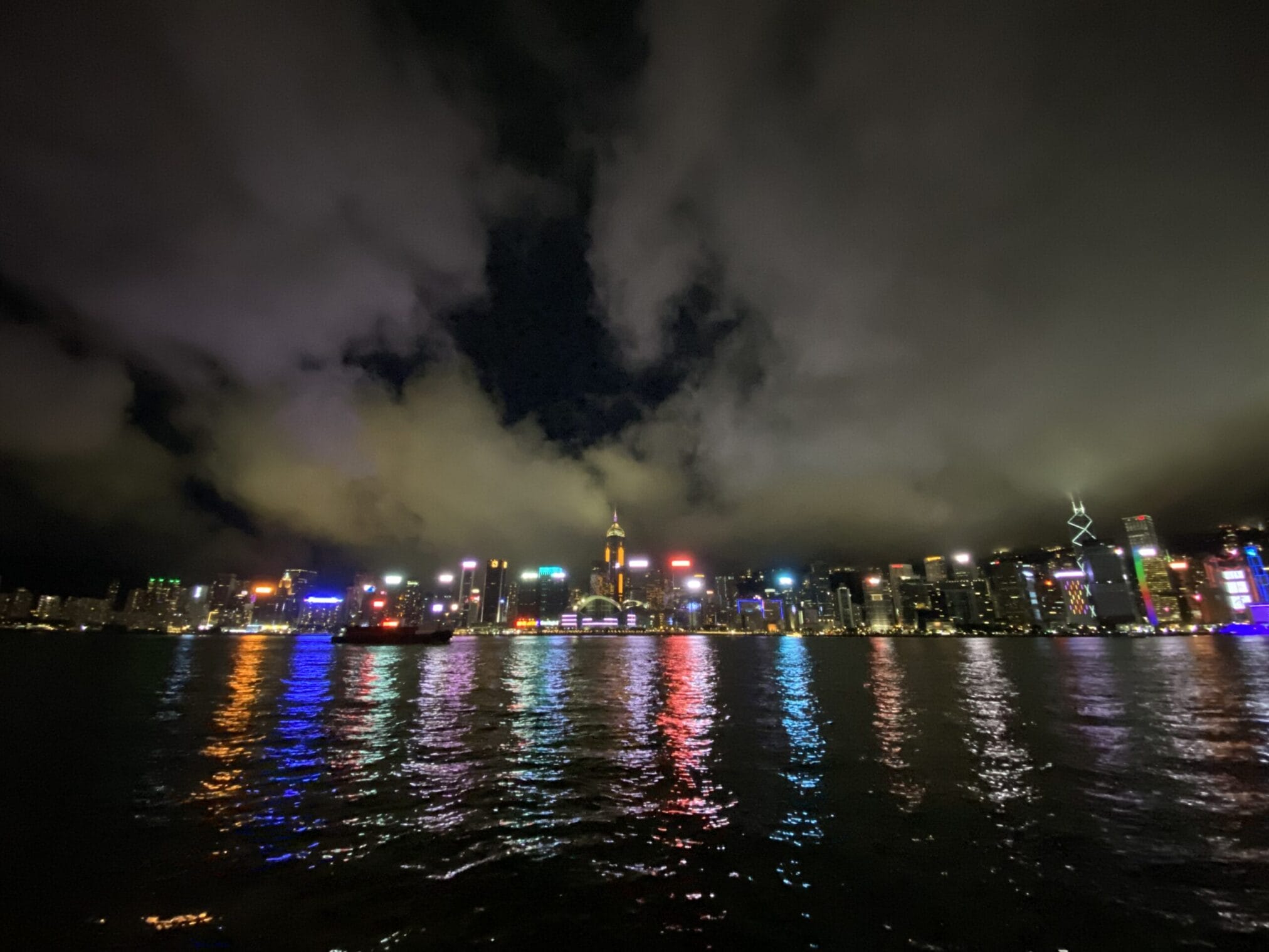 ภาพวิวเมืองฮ่องกงยามค่ำคืน มีแสงไฟจากอาคารสูงสะท้อนน้ำในอ่าวและท้องฟ้ามีเมฆหนา Hong Kong city night skyline with skyscraper lights reflecting on the harbor and cloudy sky