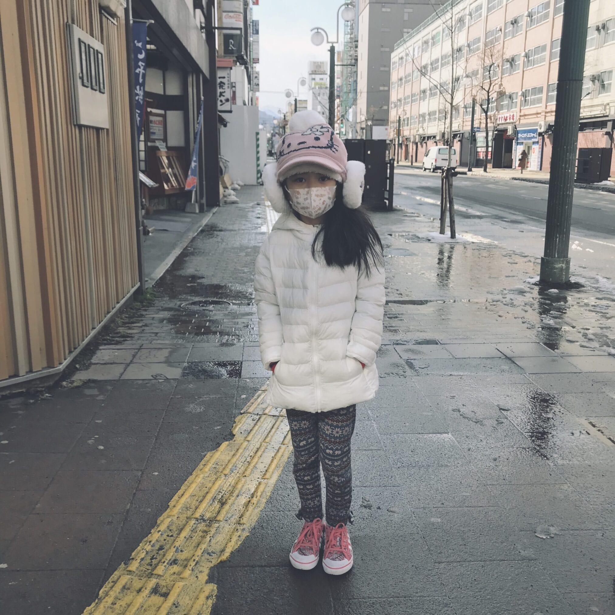 เด็กหญิงใส่เสื้อโค้ทสีขาว หมวก และหน้ากาก ยืนอยู่บนทางเท้าในฮอกไกโด