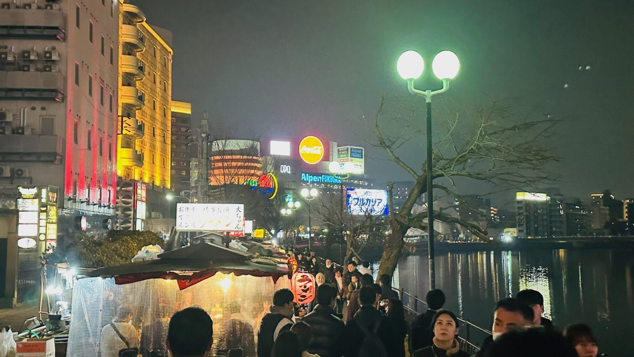 ฟุกุโอกะยามค่ำคืน มีร้านอาหารริมน้ำและป้ายโฆษณาต่างๆ เช่น Coca-Cola, Alpen Fukuoka