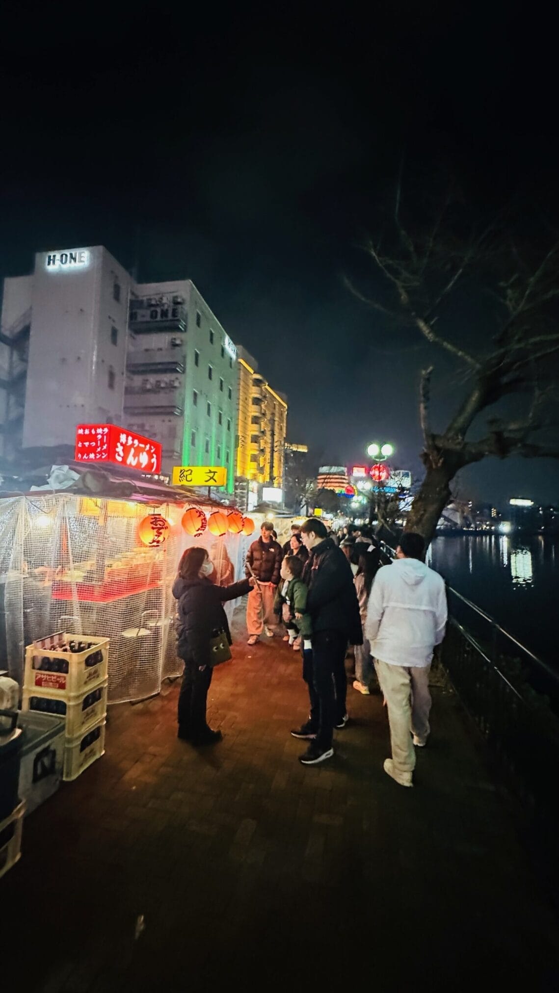 ตลาดกลางคืนริมน้ำที่ฟุกุโอกะ มีร้านแผงลอยและผู้คนสัญจรไปมา "ขับรถเที่ยวภูมิภาคคิวชู