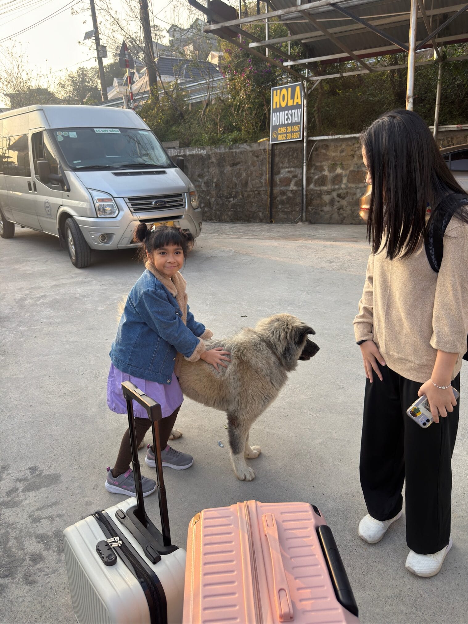 เด็กหญิงลูบหลังสุนัขหน้าโฮมสเตย์ "Hola Homestay" พร้อมกระเป๋าเดินทาง