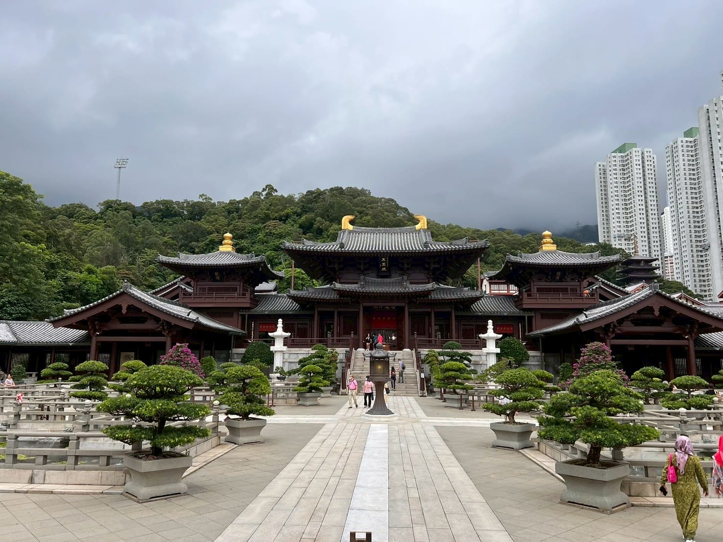 วัดสถาปัตยกรรมจีนโบราณรายล้อมด้วยต้นไม้และสวนหย่อม ตั้งอยู่ท่ามกลางภูเขาและตึกสูง Traditional Chinese temple surrounded by trees and gardens with mountains and tall buildings in the background