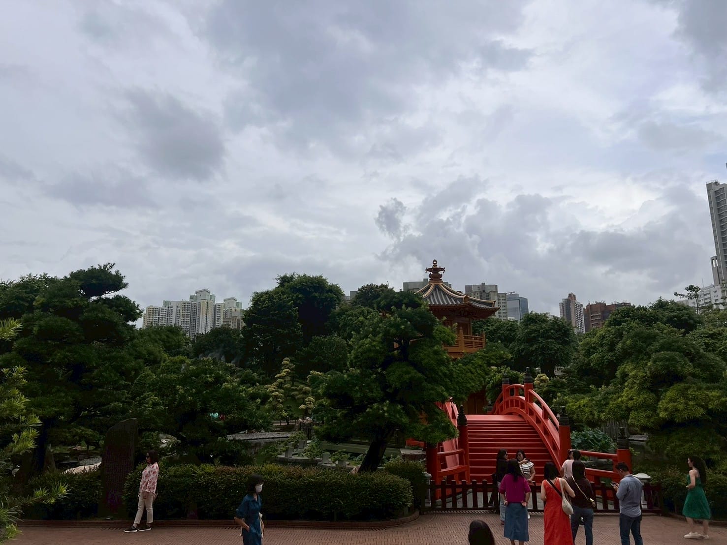 สวนญี่ปุ่นมีสะพานแดงและศาลาไม้ รายล้อมด้วยต้นไม้สีเขียวและตึกสูงในเมือง Japanese garden with red bridge and wooden pavilion surrounded by green trees and city buildings