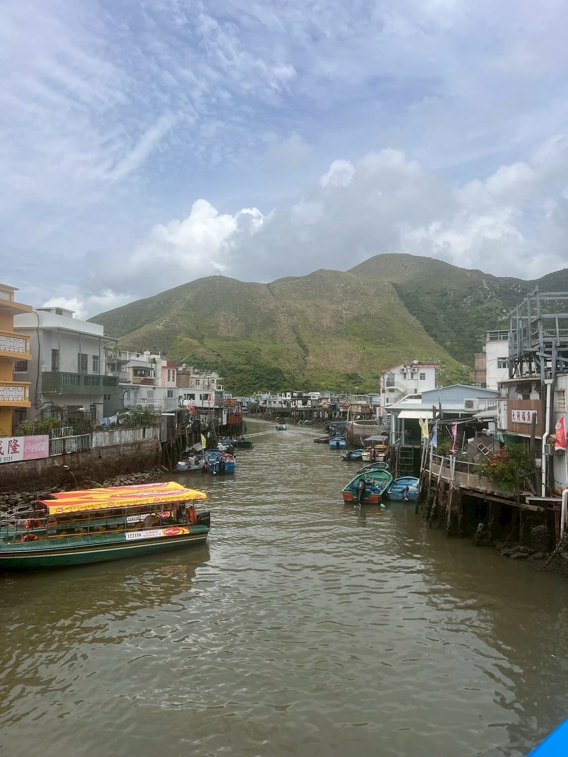 แม่น้ำลำคลองมีเรือและบ้านเรือนริมฝั่ง ภูเขาเขียวและท้องฟ้าครึ้ม River with boats and houses by the shore, green hills and cloudy sky